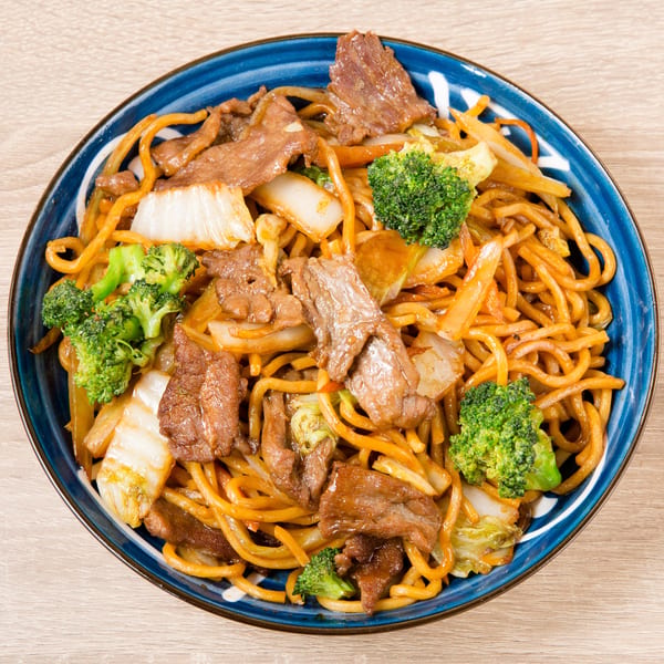 A blue bowl of beef chow mein with broccoli, cabbage, and carrots on a wooden table.