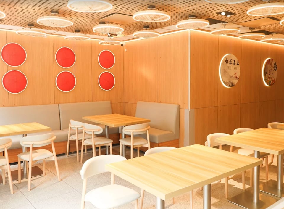 Modern restaurant interior with light wood walls, red circular decor, booth seating, and light wood tables.