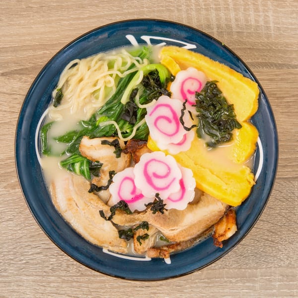 Overhead view of a ramen bowl with pork, noodles, greens, egg, and pink narutomaki.