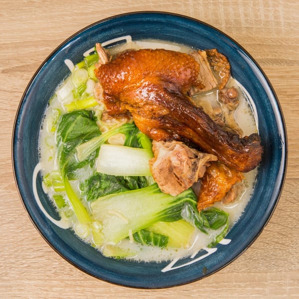 Roasted duck leg and bok choy in a creamy soup, served in a blue bowl on a wooden table.