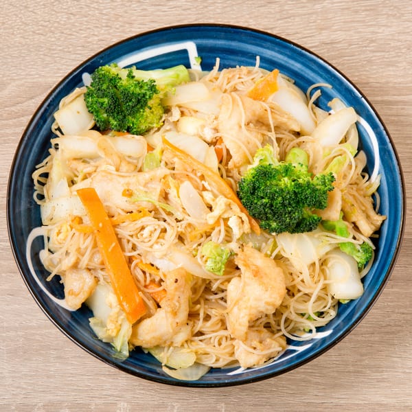 A blue bowl of Singapore noodles with chicken, broccoli, carrots, and cabbage on a wooden table.